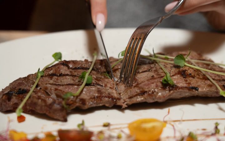 Une personne découpe une pièce de viande grillée sur une assiette blanche avec un couteau et une fourchette